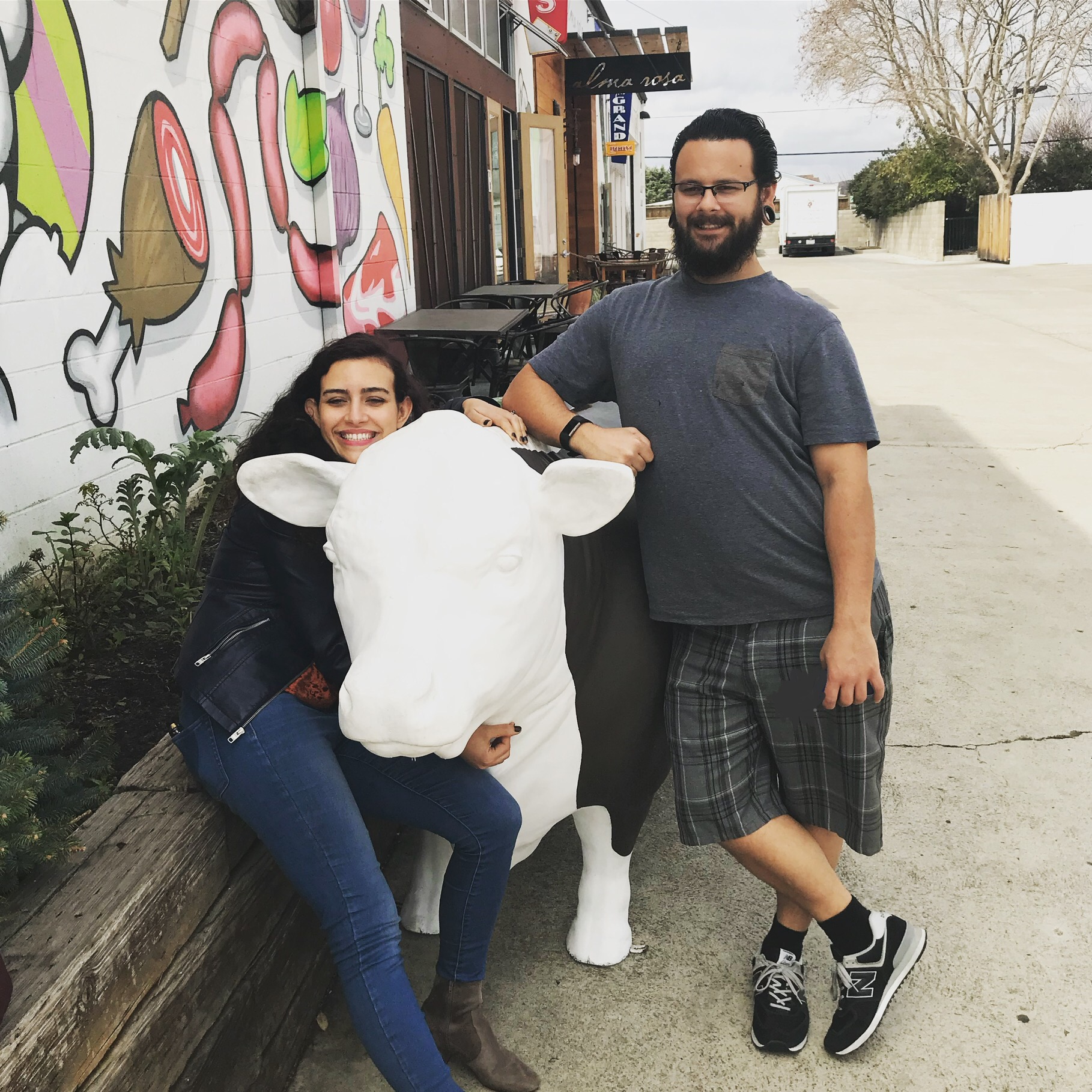 Lauren and Kori in front of Industrial Eats, posing with a cow sculpture. Graffiti of different foods is painted on the wall beside them.